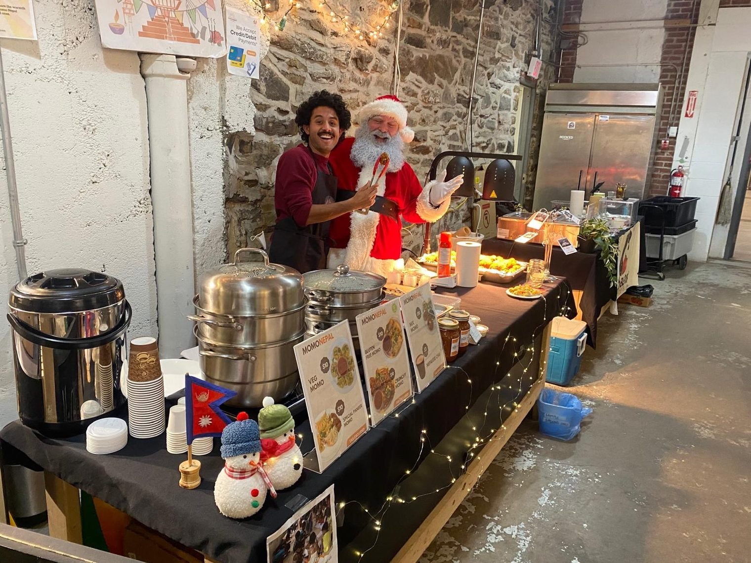 A staff member with Santa at the MomoNepal stall at Halifax Brewery Market, with the menu board and steamers on the table